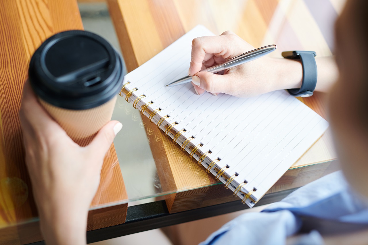 girl writing on a notebook while holding a cup of coffee perched on top of a table made of wood and glass